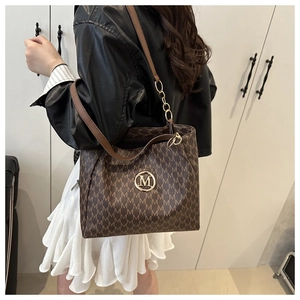 Detail of a brown tote bag with M-pattern print and golden metal logo on shoulder strap
