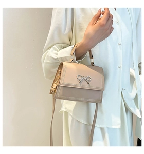 Elegante bolso de hombro beige con lazo decorativo de metal sostenuto en la mano.