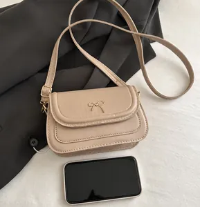 Small beige crossbody bag with gold bow detail compared to a smartphone for size