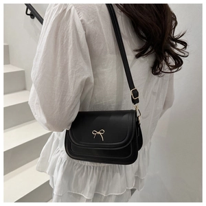 Sac à bandoulière noir minimaliste avec nœud doré porté par un mannequin en haut blanc.