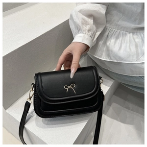 Sac à bandoulière noir minimaliste avec décoration de nœud doré sur le rabat avant.