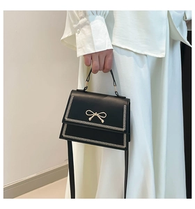 Elegante bolso bandolera negro con lazo dorado llevado por una modelo vestida de blanco.