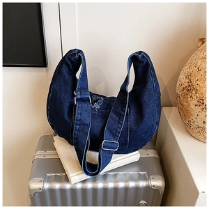 Close-up of dark blue denim crescent bag with raw edges and adjustable strap on a suitcase