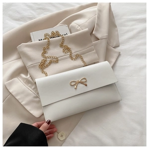 Close-up of white envelope clutch with gold bow clasp and metal chain on a beige blazer