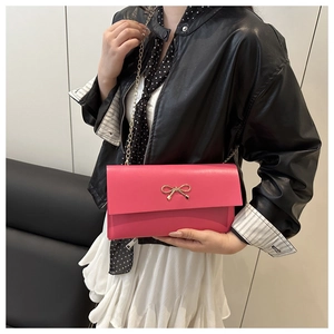 Rose pink envelope crossbody bag with gold bow detail worn by a model on black sofa