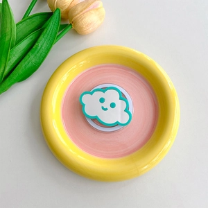 Cloud-shaped mirror phone grip with green smile face on a decorative ceramic plate