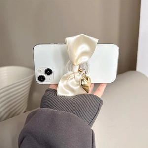 Detail view of a cream satin bow phone grip with a gold heart pendant on a transparent base