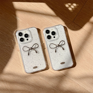Custodia iPhone glitterata con fiocco metallico 3D su superficie in legno e ombre lievi