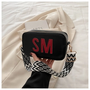 Gros plan du sac bandoulière noir avec lettre SM rouge et large sangle à motifs