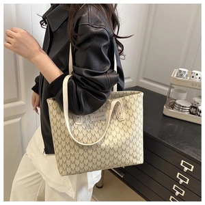 Large beige tote bag with M-pattern print and shoulder strap worn with leather jacket