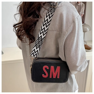 Détail d'un sac bandoulière noir avec sangle géométrique et patch lettre SM rouge