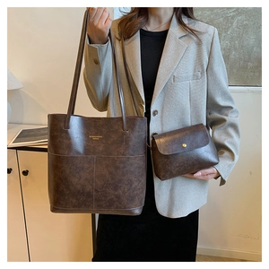 Large dark brown leather tote bag and clutch set worn by a model in a modern home setting
