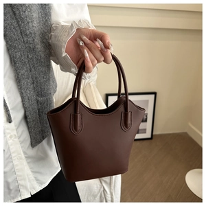 Detail of a minimalist dark brown leather bucket bag shown held by a model in a warm setting