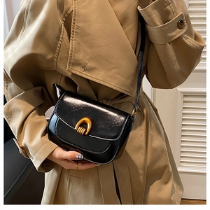 Detail of a black glossy leather saddle bag with a golden U-shaped clasp on a trench coat