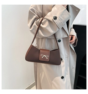 Detail of a brown leather baguette bag featuring a gold bow buckle on a beige trench coat