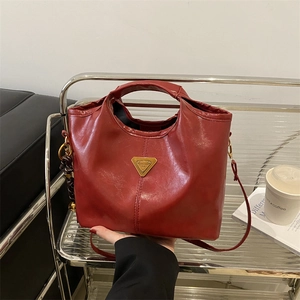 Deep red oily leather tote bag with gold triangle logo and braided charm on a metal shelf