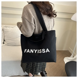 Minimalist black canvas tote bag with FANYISSA print shown in a cozy home bedroom setting