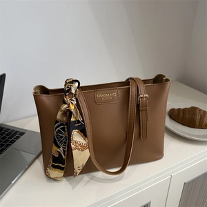 Brown leather tote bag with stylish printed scarf and fashion label on clean white desk