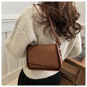 Brown leather crossbody bag with woven texture flap shown on shoulder in a chic interior