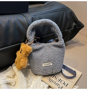 Detail of grey plush bucket bag with fuzzy bear charm and woven strap on a soft surface