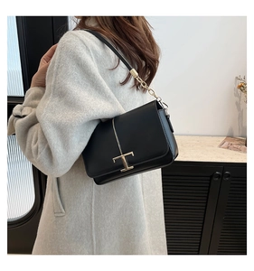 Détail d'un sac bandoulière en cuir noir avec boucle en T dorée porté sur manteau gris