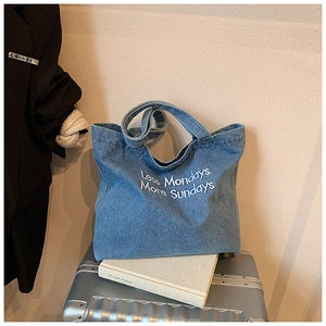 Detail of blue denim tote bag with Less Mondays More Sundays white embroidery on metallic suitcase