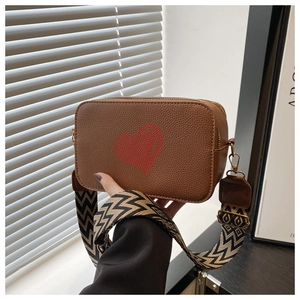 Brown camera bag with red heart print and striped wide strap held in hand