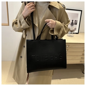 Detail of black faux leather tote bag with embossed TOTE BAG lettering on beige trench coat