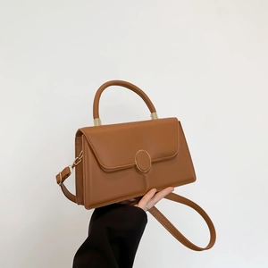 Structured caramel brown leather handbag with gold buckle held against a neutral wall
