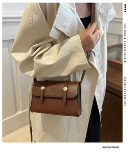 Brown leather crossbody bag with gold metal buckles worn over a beige trench coat