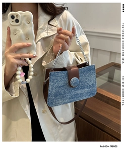 Detail of blue denim tote bag with silver chain handle and leather strap on a beige trench coat