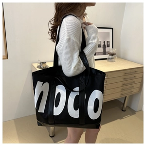 Close-up of a large black noco canvas tote bag worn by a model in a cozy interior setting