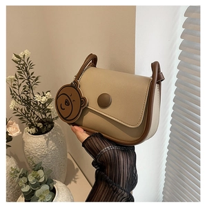 Détail d'un sac selle beige avec bordure marron et breloque ours dans un décor de vie