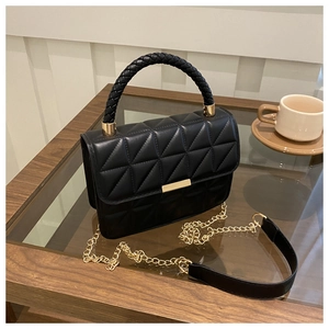 Detail of black quilted crossbody bag with woven handle and gold hardware on a glass table