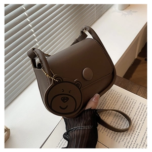 Détail d'un sac besace marron mat avec boucle ronde et breloque ours sous une douce lumière