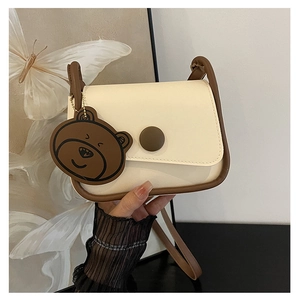 Détail du sac selle crème avec liseré marron et pendentif ours mignon tenu à la main