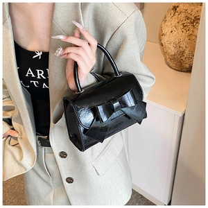 Detail of a black patent leather handbag with a large bow held by a model in a blazer