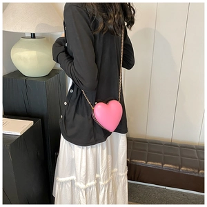 Pink heart-shaped crossbody bag with gold chain strap worn by a model in a minimal setting