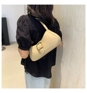 Close-up of beige glossy hobo bag with a large gold buckle worn by a model in a room