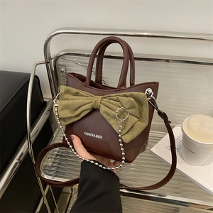 Brown leather shoulder bag with green bow and silver chain on a modern metal stand