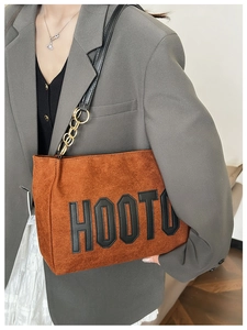 Détail du cabas en daim carmél avec écusson HOOTU et chaîne dorée sur un blazer gris