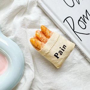 Detail shot of the baguette-shaped silicone case inside a beige paper bag on white linen