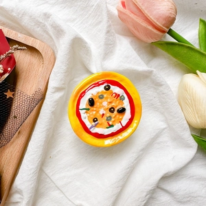 Detail view of a 3D pizza-shaped phone grip on white textured fabric background