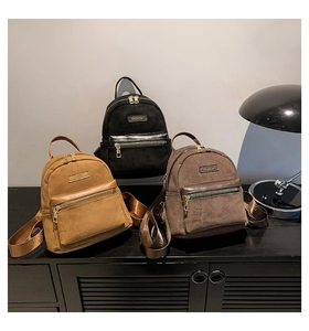 Three trendy velvet backpacks in black, brown, and camel on a display shelf with metal labels