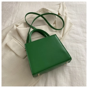 Détail du sac carré vert avec pieds de protection dorés sur tissu blanc texturé