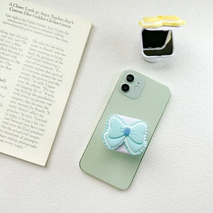Close-up of a blue bow collapsible phone grip with a yellow makeup mirror on a white table