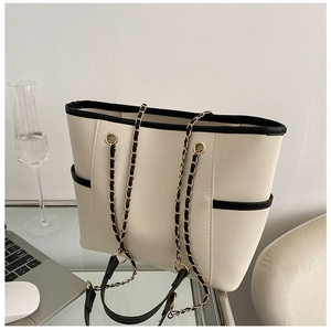 Detail of cream tote bag with black trim, gold chain strap, and side pocket on desk