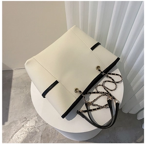 Close-up of cream tote bag with black trim and gold chain shoulder strap on a white podium