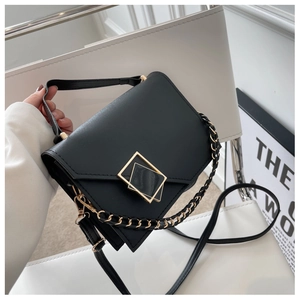 Detail of black structured handbag with gold square buckle and woven chain on a white chair