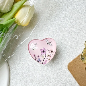 Detail view of a pink heart-shaped phone grip featuring floral and dragonfly art patterns